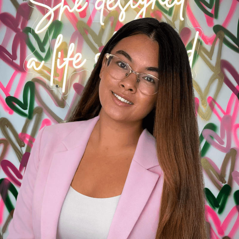 Woman wearing glasses and a pink blazer in front of a colorful wall with text.